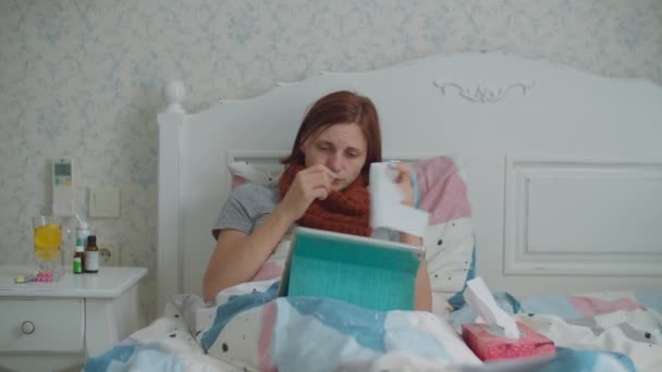 Beteg fiatal nő, aki tabletet használ az ágyban fekve. Egy nő meleg sállal a nyakán köhög és orrát fújja, miközben beszél az orvossal online. Influenza és más vírusbetegségek. Online orvosi konzultáció. — Stock videók