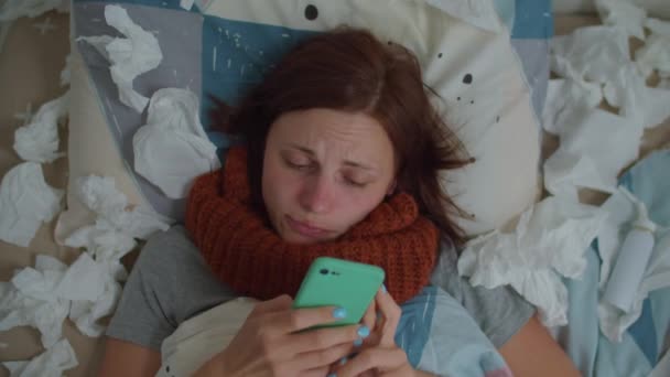 Sick young 30s woman uses mobile phone and blows her nose laying in bed with lots of napkins around. Woman with running nose suffers from virus disease at home. — Stock Video