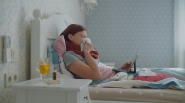 Yatakta tablet bilgisayar kullanan hasta genç bir kadın. Boynunda eşarp olan, öksüren ve burnunu silen bir kadın. Grip ve diğer solunum hastalıkları.