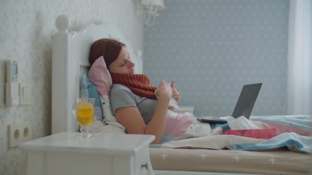 Sjuk ung kvinna som ligger i sängen och tittar på laptop på nätet. Kvinna med varm halsduk på halsen hostar, blåser näsan och dricker hett te med citron. Influensa och andra luftvägssjukdomar i hemmet. — Stockvideo