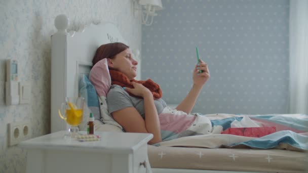 Zieke jonge vrouw met een mobiele telefoon in bed. Vrouw met warme sjaal op de nek hoest en blaast neus tijdens het praten met arts online. Griep en andere ademhalingsziekten. Online doktersconsult. — Stockvideo