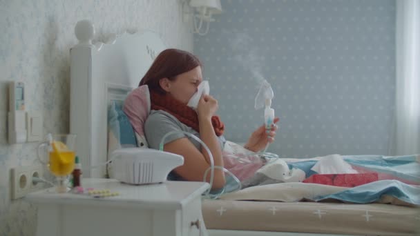 30代の若い女性は、ナプキンで鼻を吹いて、自宅でインフルエンザや呼吸器ウイルスなどの病気の治療のために吸入器を使用しています。女性の治療咳で薬吸入器マシンはベッドに横たわっています. — ストック動画