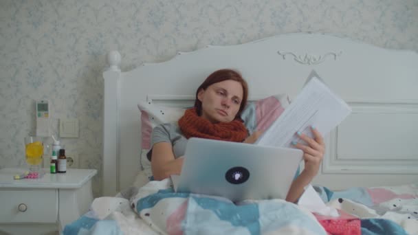 Sick young woman working from home laying in bed with documents and laptop. Woman with warm scarf on neck coughs, blows her nose and looking at documents. Flu and other respiratory diseases at home. — Stock Video