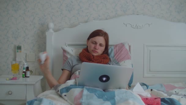 Zieke jonge vrouw die thuis met een laptop in bed ligt. Vrouw met warme sjaal op de nek hoest, blaast haar neus en kijkt naar de computer. Griep en andere virusziekten thuis. — Stockvideo