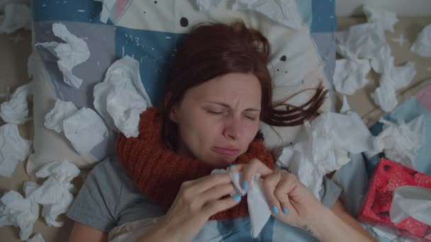 Sick young 30s woman blows her nose laying in bed with lots of napkins around. Woman with running nose suffers from virus disease at home. Top view. — Stock Video