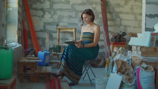 Ung vuxen kvinna i klänning tittar igenom ritningar av hus renovering på surfplatta dator på byggarbetsplatsen. Kvinnlig dröm om ny inredning. Person som gör bilder på ny lägenhet. — Stockvideo
