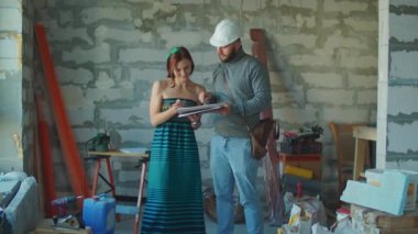 Beyaz kasketli bir erkek ustabaşı. Belgeler ve apartman onarımı planları gösteriyor. Ev onarımı için tamirciyle evrakları imzalayan emlakçı kadın..