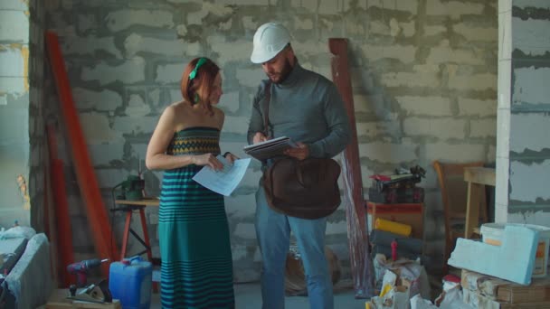 Mujer adulta joven comprueba un plan de construcción con capataz en casco de seguridad blanco. Señora hablando con el reparador y mirando los documentos en el sitio de construcción. — Vídeos de Stock