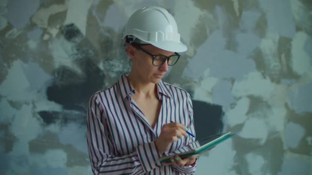 Trabajadora joven en hardhat blanco y gafas de pie con tableta en las manos. Capataz revisando el proceso de renovación de la casa. Mujer en casco blanco sonriendo mirando a la cámara. — Vídeos de Stock