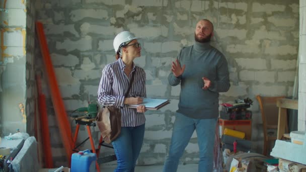 Joven barbudo discutiendo un plan de construcción con una capataz en un sombrero blanco. Hombre y mujer hablando en el sitio de construcción, sosteniendo planos. — Vídeos de Stock