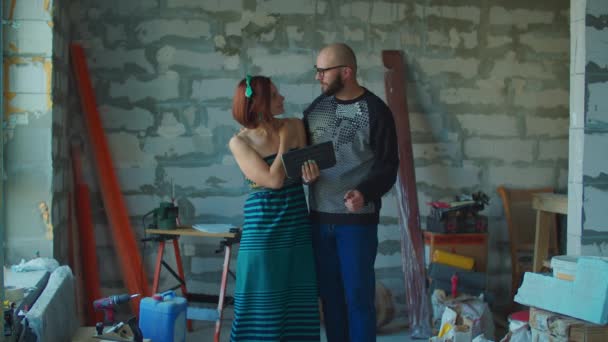 Jong opgewonden koppel plannen van hun nieuwe huis renovatie kijken naar blauwdrukken op tablet computer staan op de bouwplaats. Gelukkig millennial familie knuffelen. — Stockvideo