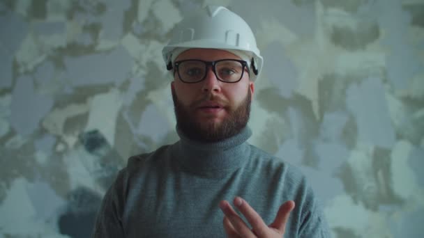 Een man met een baard in de 30 met een witte hoed en een bril die naar de camera kijkt. Foreman legt woningrenovatieproces uit. — Stockvideo