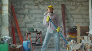 Beyaz koruyucu takım elbiseli genç, komik bir kadın işçi inşaat alanındaki mikrofon gibi patenle dans edip şarkı söylüyor..