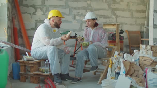 Dos pintores almorzando en la obra. Hombre y mujer en sombreros sentados en los materiales de construcción y hablando mientras come. — Vídeos de Stock