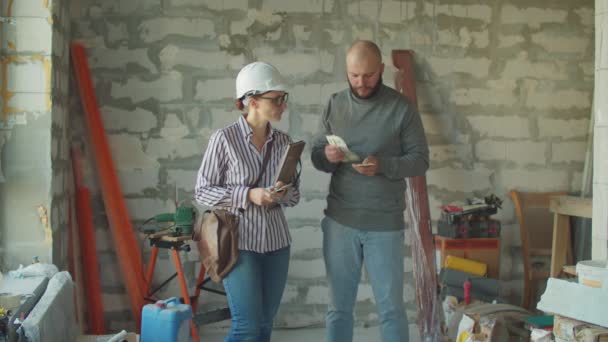 Junger bärtiger Mann gibt Frau mit weißem Bollenhut Geld. Männlicher Grundstückseigentümer zahlt für Haussanierung auf Baustelle. — Stockvideo