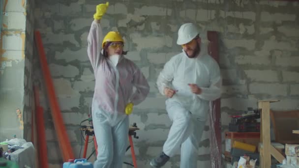 Молода смішна жінка і чоловік-працівник в білих захисних костюмах танцює на будівельному майданчику. Позитивний процес ремонту будинку. Люди в захисних капелюхах . — стокове відео