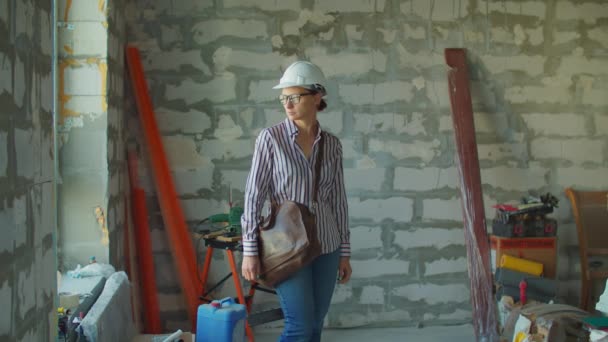 Trabajadora joven en hardhat blanco de pie en el sitio de construcción. Capataz revisando el proceso de renovación de la casa. Mujer con casco blanco dentro. Señal de caja "Mezcla de polvo termoendurecido" — Vídeos de Stock