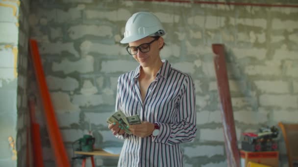 Fiatal, mosolygós, fehér keménykalapos női munkás, aki az építkezésen áll. Foreman pénzt tart magánál a ház felújítására. "Hőre keményedő porkeverék" doboztábla — Stock videók