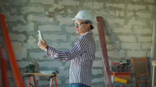 Egy fehér keménykalapos fiatal nő áll az építkezésen mobiltelefonnal a kezében. Foreman videohívással beszél az ügyféllel a házfelújítási folyamatról. "Hőre keményedő porkeverék" doboztábla — Stock videók