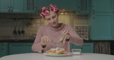 Kafasına pembe bigudiler serpiştirilmiş, ev yapımı tarçın ve şeker serpiştirilmiş genç ve komik bir kadın. Evde tek başına yemek yapmak..