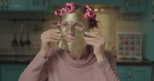 Mujer de 30 años con rizadores rosados en la cabeza aplicando máscara cosmética de oro en su cara. Joven mujer adulta disfruta de la rutina de belleza en casa. De cerca.. — Vídeo de stock