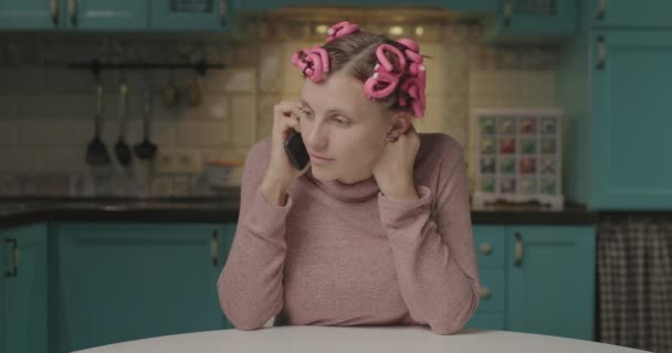 Молодая 30-летняя женщина с бигудями на голове разговаривает по мобильному телефону дома. Забавная женщина болтает с подругой за камерой, сидя на кухне. — стоковое видео