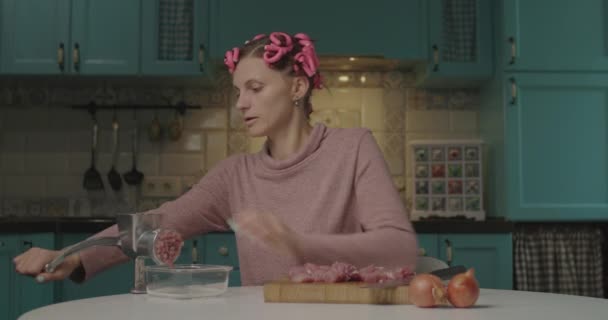 30-jährige junge Frau mit Lockenwicklern auf dem Kopf, die in der Küche Fleisch im Fleischwolf kocht. Hausfrau kocht allein zu Hause. — Stockvideo