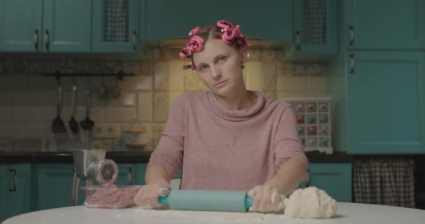 Eine junge Frau in den Dreißigern rollt den Teig mit einem Nudelholz aus und schaut in die Kamera, die in der Küche sitzt. Hausfrau mit Lockenwickler kocht allein zu Hause. — Stockvideo