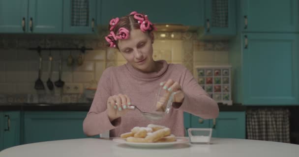 Mulher de aparência engraçada nova com rolos rosa na cabeça polvilha eclairs caseiros com canela e açúcar. Cozinhar sozinho em casa. — Vídeo de Stock