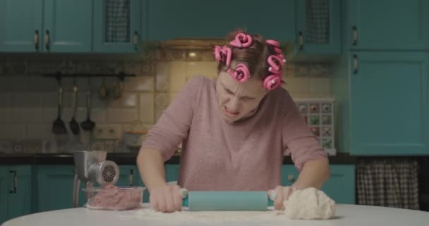 Молода розлючена доросла жінка викидає тісто з прокатним штифтом, сидячи на кухні. Домогосподарка з праскою для волосся втомилася готувати вдома наодинці. Жінка дивиться сердито на камеру . — стокове відео