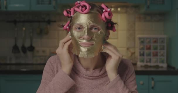 30s woman with pink curlers on head applying gold cosmetic mask on her face. Young adult female enjoys home beauty routine. Close up. — Stock Video