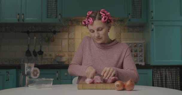 Grappige jonge vrouw met haarkrulspelden op het hoofd vlees snijden met mes zittend in de keuken. Huisvrouw koken vlees in gehakt. — Stockvideo