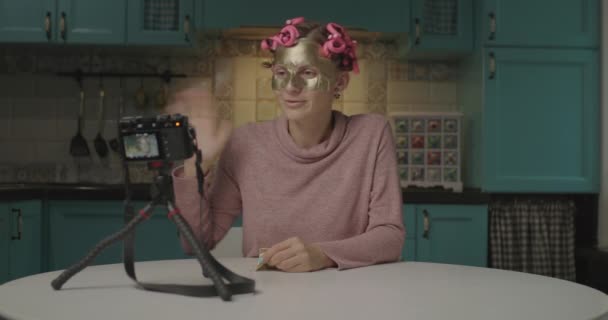 Joven mujer de 30 años grabando divertido video de la aplicación de la máscara cosmética de la cara en casa. Divertida blogger femenina con rizadores de pelo haciendo video tutorial de belleza para el canal de redes sociales. — Vídeo de stock