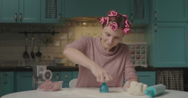 Ung vuxen kvinna gör hål i degen sittandes i köket. Le hemmafru med hår locktång matlagning hemma ensam. Dough rolig mask. — Stockvideo