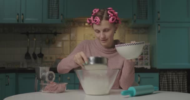 Giovane donna divertente impastare pasta seduta in cucina. Casalinga con bigodini sulla testa mescolando farina con acqua per fare pasta per gnocchi a casa da sola. — Video Stock