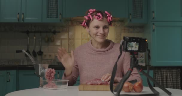 女性フードブロガーは、ミンサーで肉を調理し、キッチンに座ってカメラにビデオを記録します.髪の毛のカーラーを持つ主婦が自宅で調理. — ストック動画