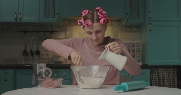 Grappige jonge vrouw die deeg kneedt zittend in de keuken. Huisvrouw met haarkrulspelden op het hoofd bloem mengen met water om deeg te maken voor dumplings alleen thuis. — Stockvideo