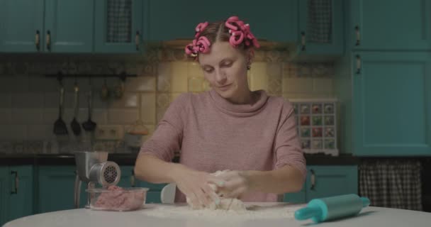 Grappige jonge vrouw die deeg kneedt zittend in de keuken. Huisvrouw met haarkrulspelden op het hoofd bloem mengen met water om deeg te maken voor dumplings alleen thuis. — Stockvideo