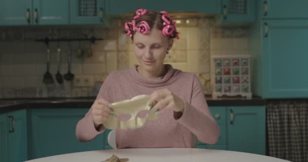 Een vrouw uit de 30 met roze krulspelden op haar hoofd en een gouden cosmetisch masker op haar gezicht. jong volwassen vrouw geniet thuis schoonheid routine op blauw keuken. — Stockvideo