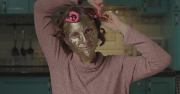 Jonge vrouw van in de 30 met een gouden cosmetisch masker op haar gezicht die haar krultang afdoet terwijl ze naar de camera kijkt. jong volwassen vrouw maken haar haar krullen. — Stockvideo