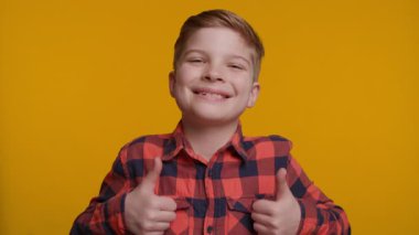 Neşeli bir çocuk doğrudan kameraya bakarken başparmağını kaldırıyor. Parlak sarı arka plan, onun kendine güvenen ve mutlu ifadesine enerji ve pozitiflik katıyor..