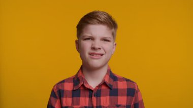 Çocuğun buruşuk bir yüzü var, rahatsızlık ya da iğrenme belirtileri gösteriyor. Arkaplan koyu sarı bir renktir. Kısa, düzgün taranmış saçları ve açık tenli genç bir çocuk kırmızı ve siyah bir ekose gömlek giyiyor.. 