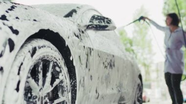 Bir kadın açık havada araba yıkamada arabasını yıkamak için hortum ve sabunlu su kullanıyor. Olay yeri, sıradan bir ortamda araba bakım rutinini yakalıyor..