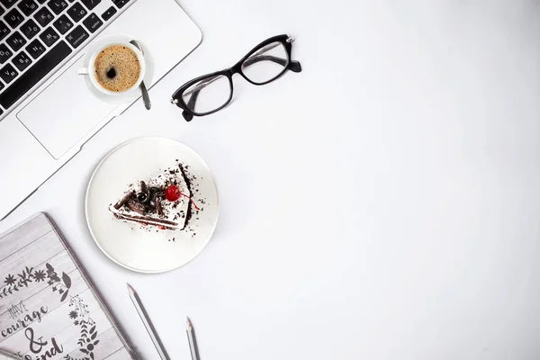 Top view of work place table, laptop with glasses notebook, cake and cup of coffee on white background