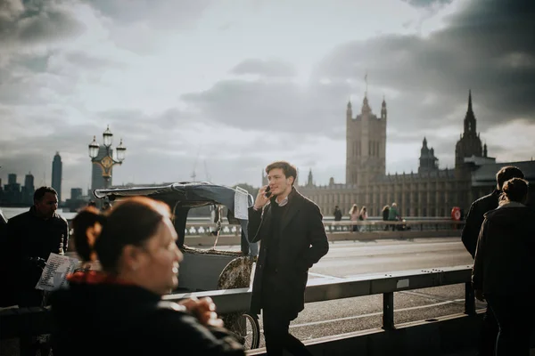 2018 日のロンドン イギリス ロンドン イングランドで バック グラウンドでウェストミン スター宮殿との電話で呼び出し中ウェストミン スター橋歩道で歩いて若い男 — ストック写真