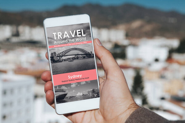 Man holding a mobile phone in the hand with travel agency website in the screen, with city background.