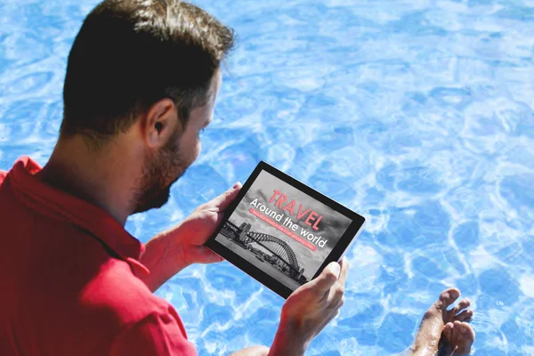 Man holding a tablet with travel agency website in the screen, while sitting on the poolside.