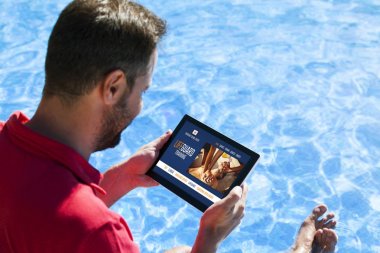 Ekranda acil servis eğitim web sitesi ile bir tablet tutan adam, yüzme havuzu kıyısında otururken.