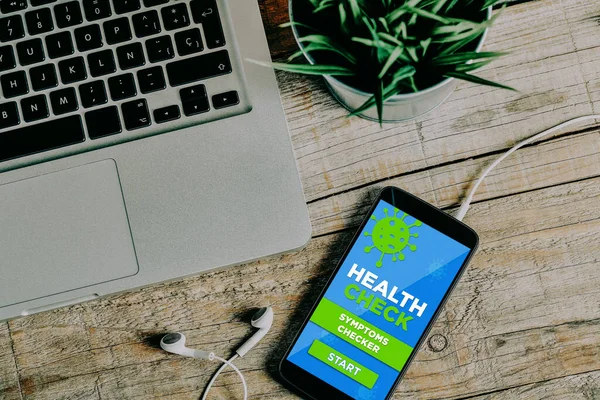 Health and symptoms checker app in a mobile phone screen placed on a wooden office desk, close to a laptop computer.