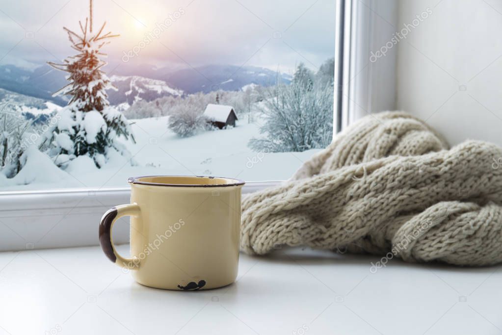 Winter background - cup with candy cane, woolen scarf and gloves on windowsill and winter scene outdoors. Still life with concept of spending winter time at cozy home with cold weather outdoors